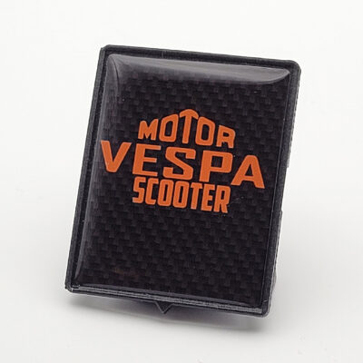 Schwarzes Vespa-Piaggio-Emblem in Carbon-Optik mit orangefarbenem „Motor Vespa Scooter“-Schriftzug.