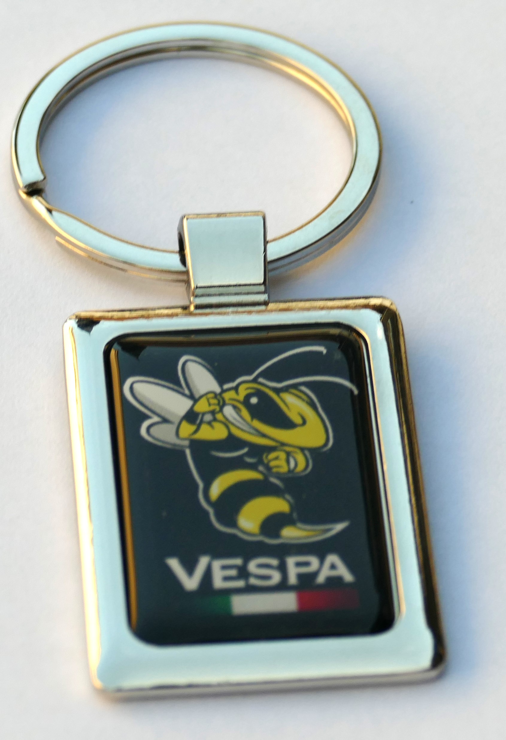 Metallischer Schlüsselanhänger mit Vespa-Motiv: gelb-schwarze Wespe auf dunklem Hintergrund, darunter der Schriftzug „VESPA“ und die italienische Flagge.